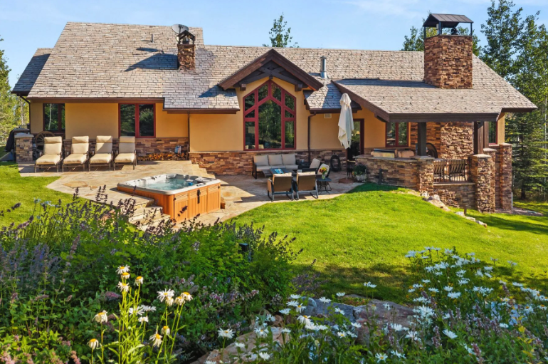 A scenic view of a modern house with stone and wood accents, featuring an outdoor hot tub and patio area, surrounded by lush greenery and colorful flowers.