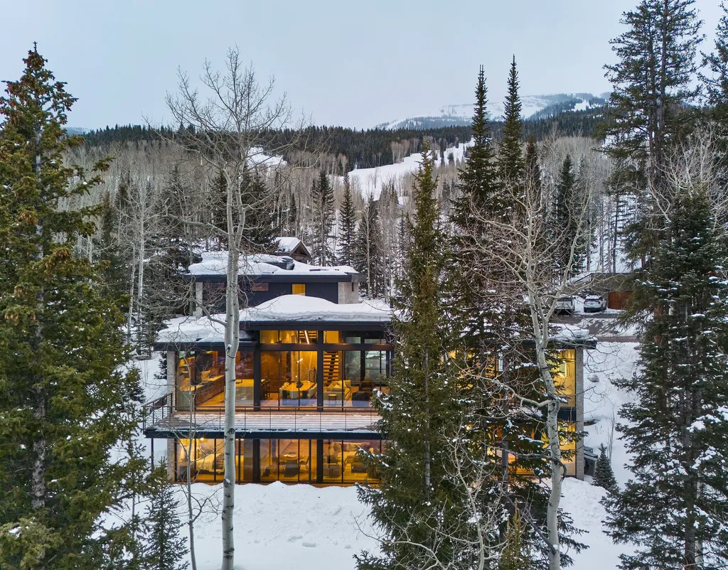 A modern house surrounded by snow-covered trees, featuring large glass windows that reflect the warm interior lighting, set against a backdrop of distant snowy hills.
