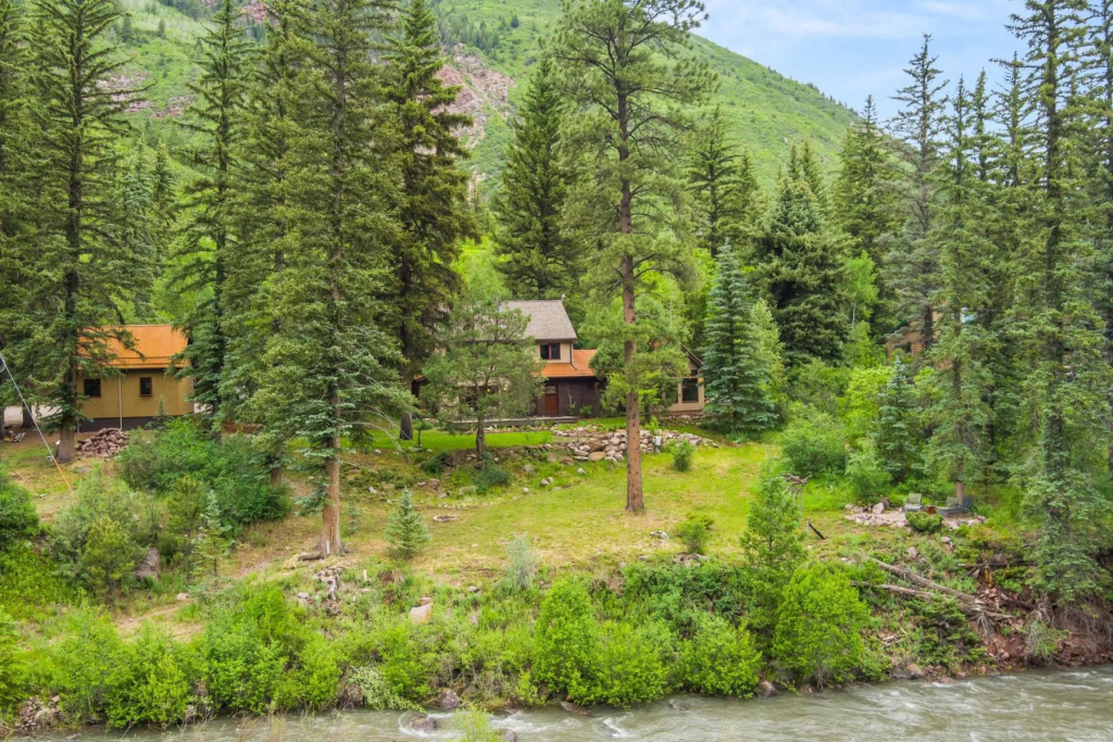 A scenic view of a cozy house nestled among tall pine trees on a green hillside, with a river flowing nearby.