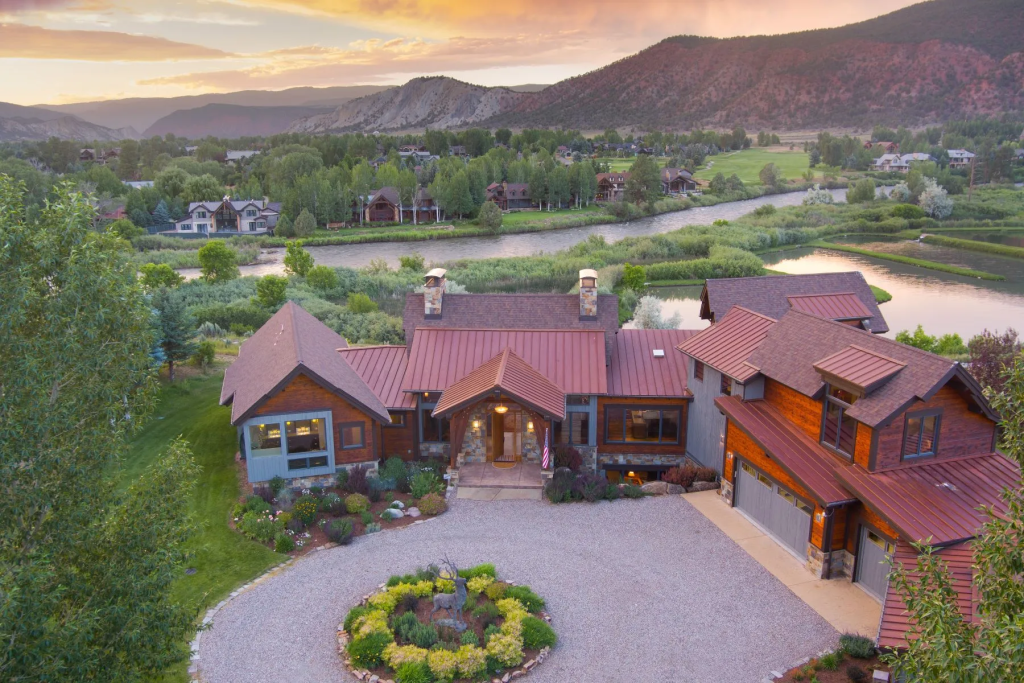 Aerial view of a modern wooden house with a red metal roof, surrounded by landscaped gardens and trees, beside a river with mountains in the background during sunset.