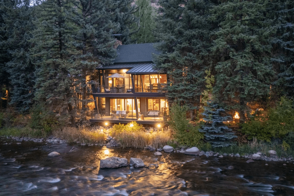A modern two-story house surrounded by tall trees, with large glass windows and a deck overlooking a river, illuminated at dusk.