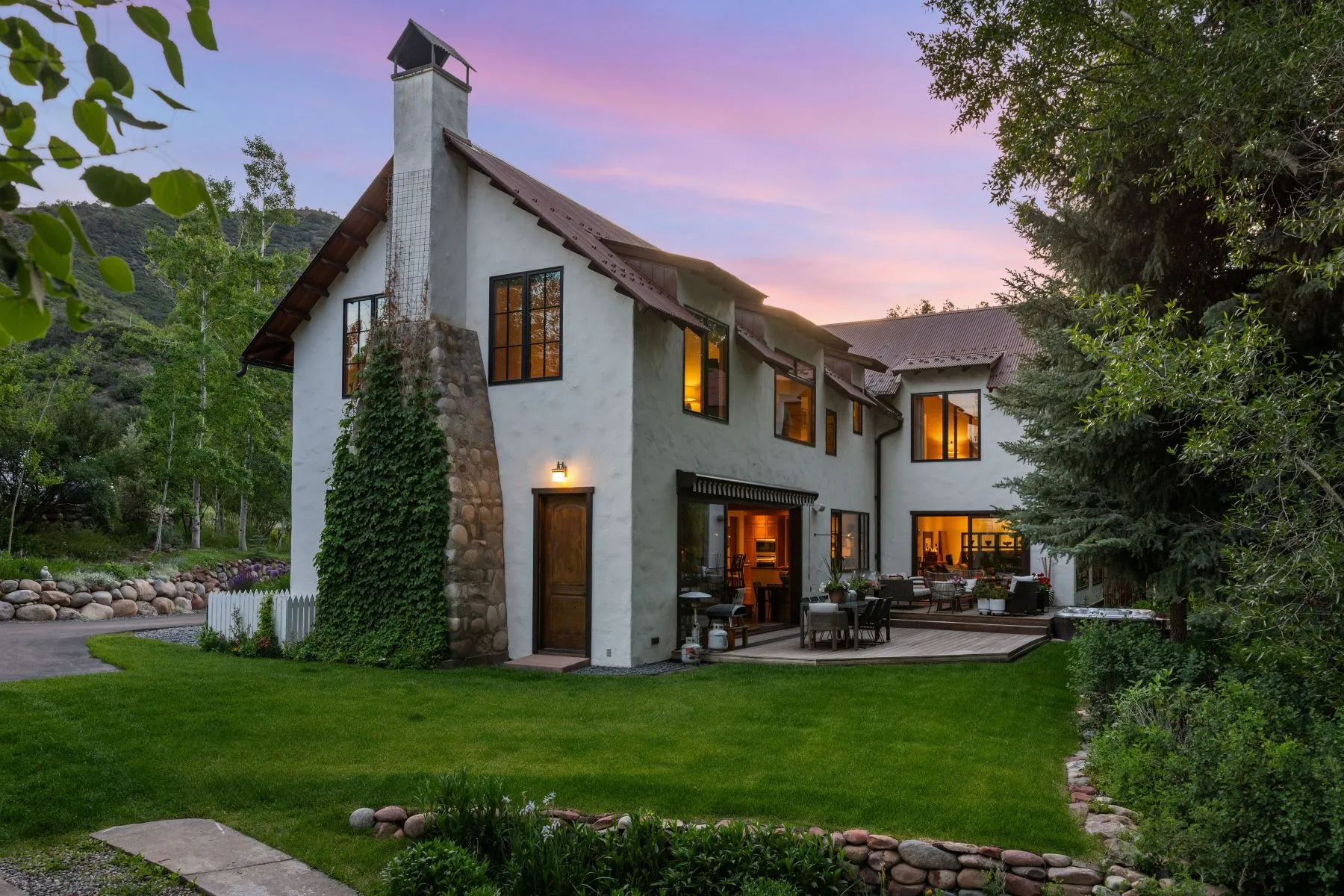 A charming two-story house with a stone chimney, surrounded by green grass and trees, featuring large windows and a wooden deck with outdoor seating, set against a colorful sunset sky.